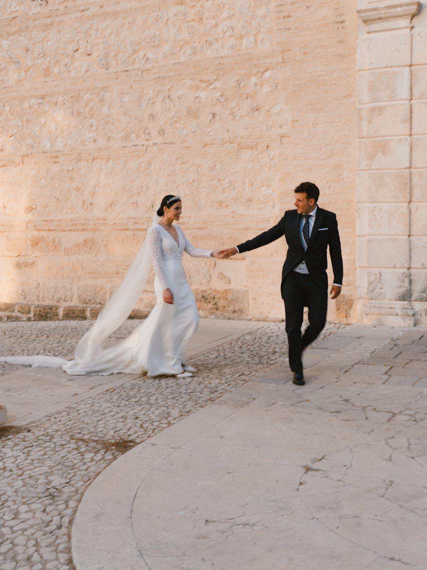 Los novios caminando de la mano junto al muro de piedra